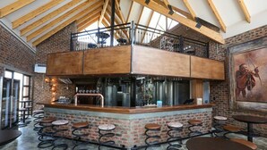 A bar area with a brick wall, wooden ceiling beams, and a mezzanine level. There are stools and tables around the bar. - Thanda Tau - Hostel (Phumelela)