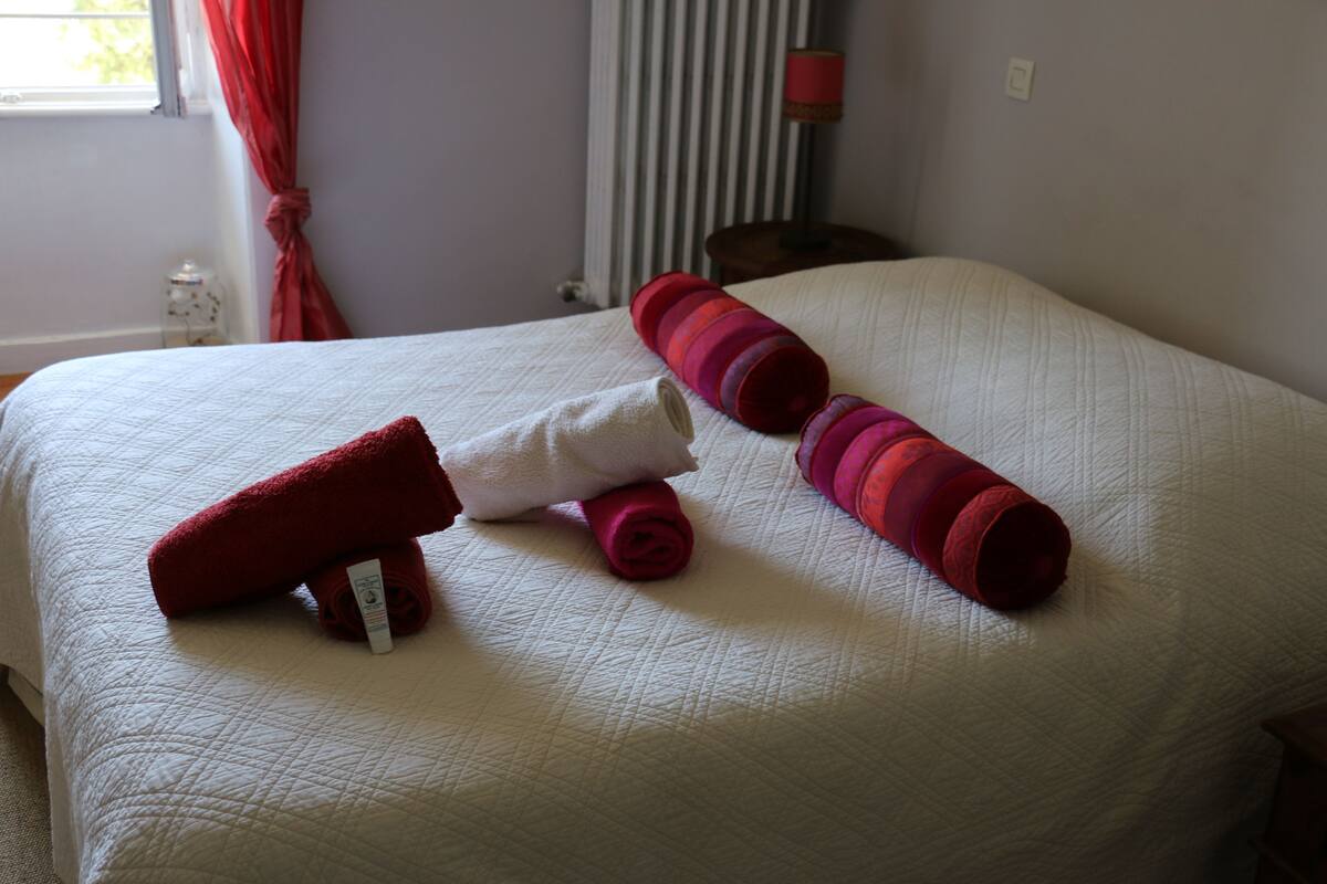 A neatly made bed with white and red linens, a small bedside table with a lamp, and a window with curtains.