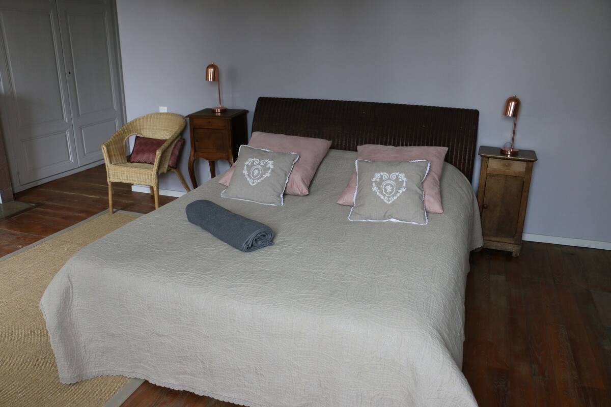 A neatly made bed with a beige quilt, two bedside lamps, and a chair with a cushion.