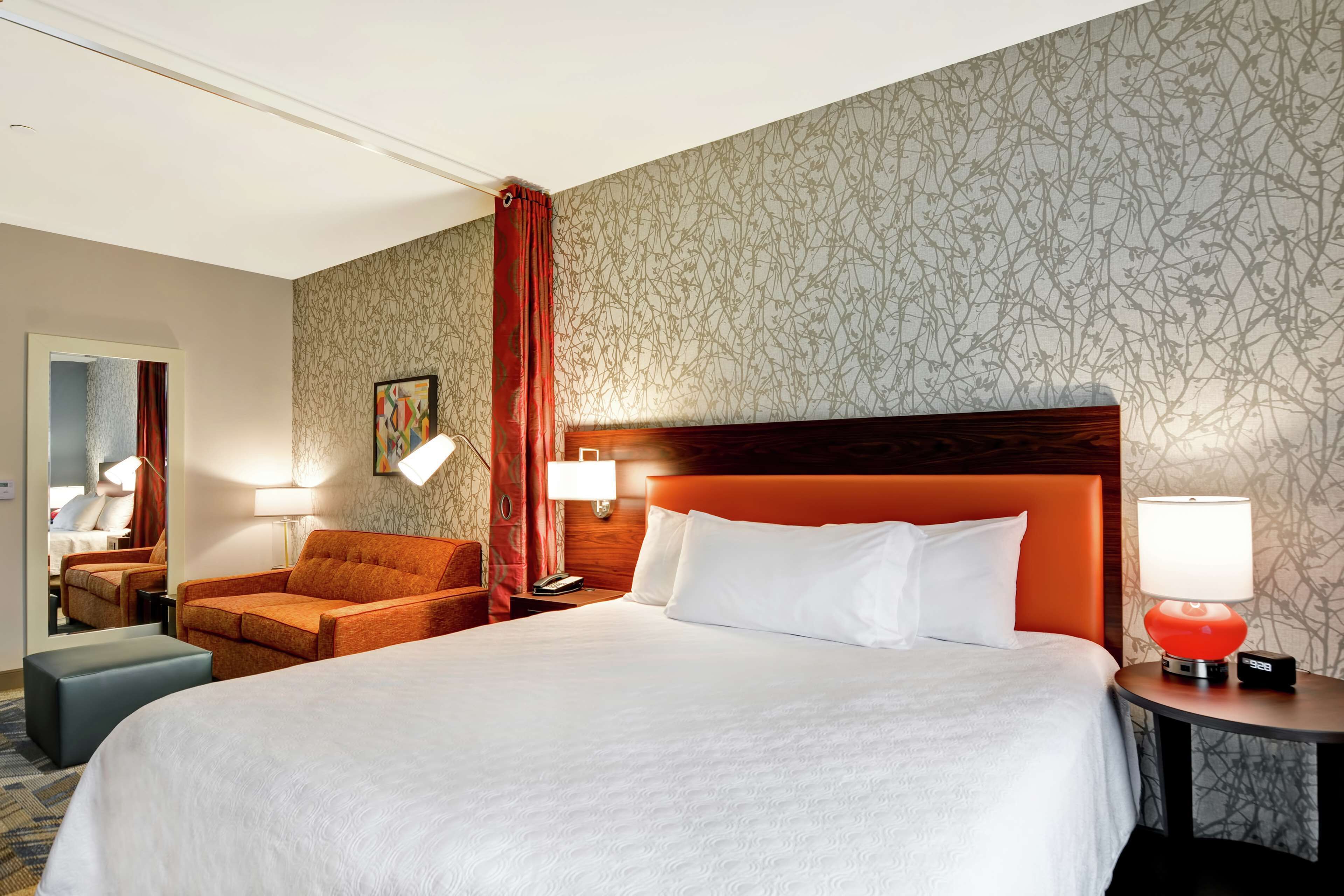 A hotel room with a large bed, a wooden headboard, a red chair, a nightstand with a lamp, and a mirror.