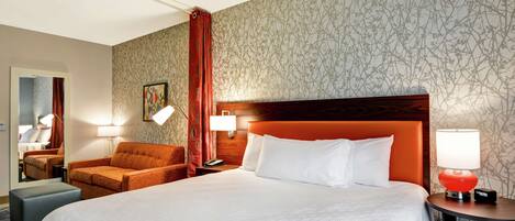 A hotel room with a large bed, a wooden headboard, a red chair, a nightstand with a lamp, and a mirror.