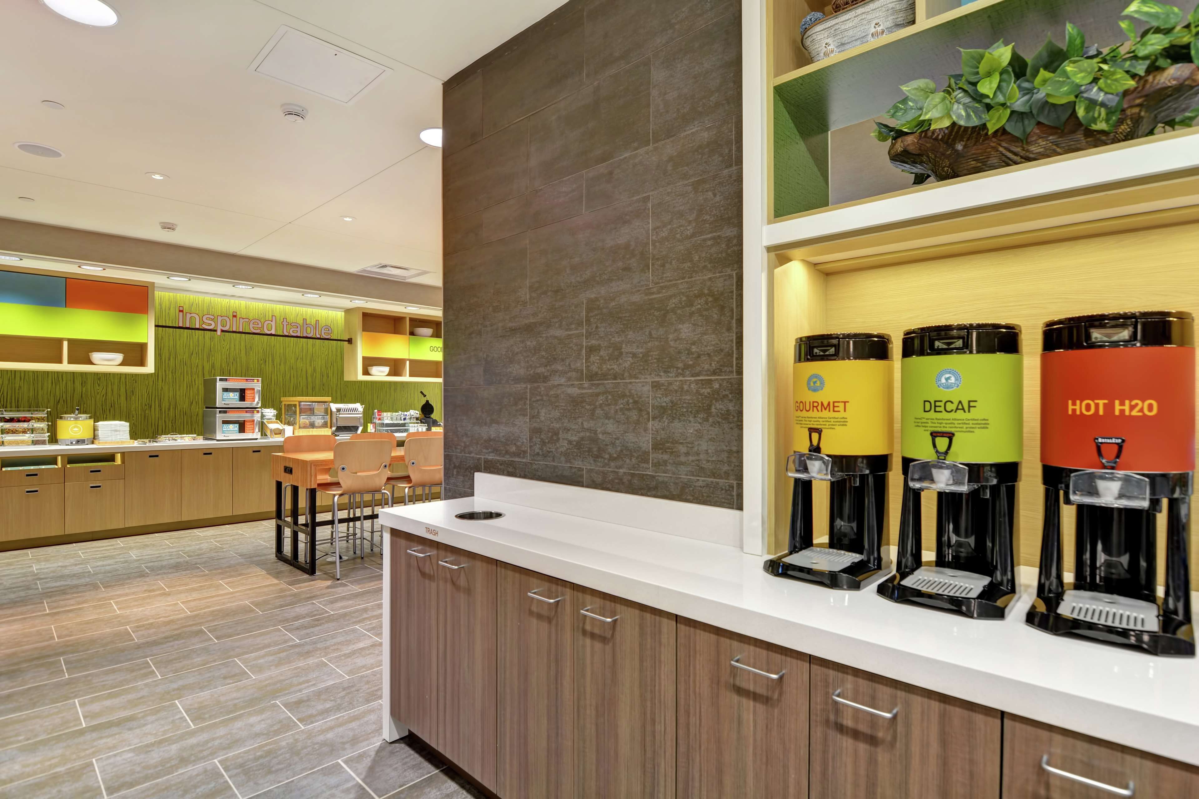 A modern kitchen area with a coffee station featuring branded dispensers and a counter with a green wall and a sign reading "inspired table".