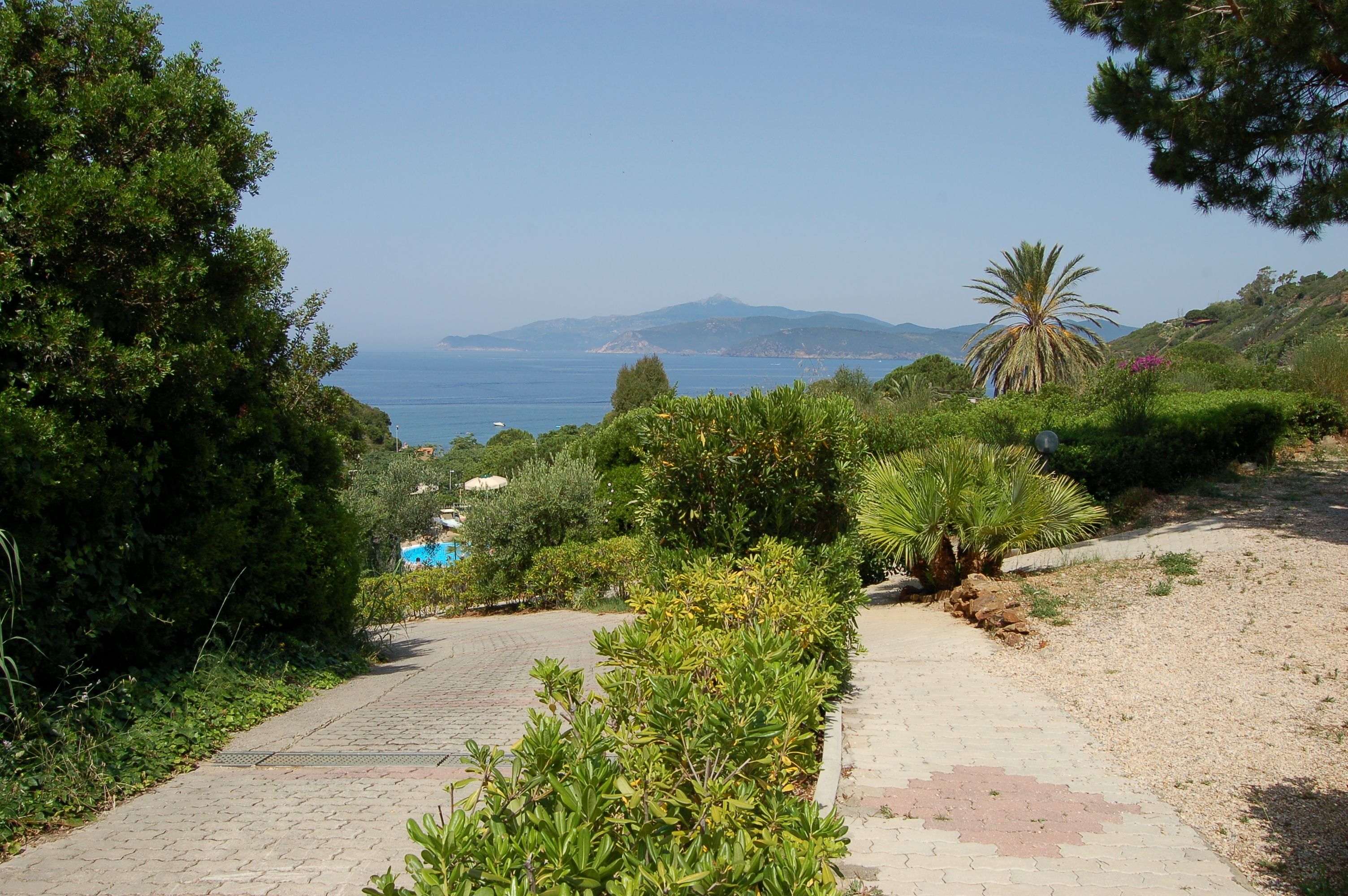 Un camino pavimentado que lleva a una playa con abundante vegetación y una vista despejada del mar y montañas distantes.