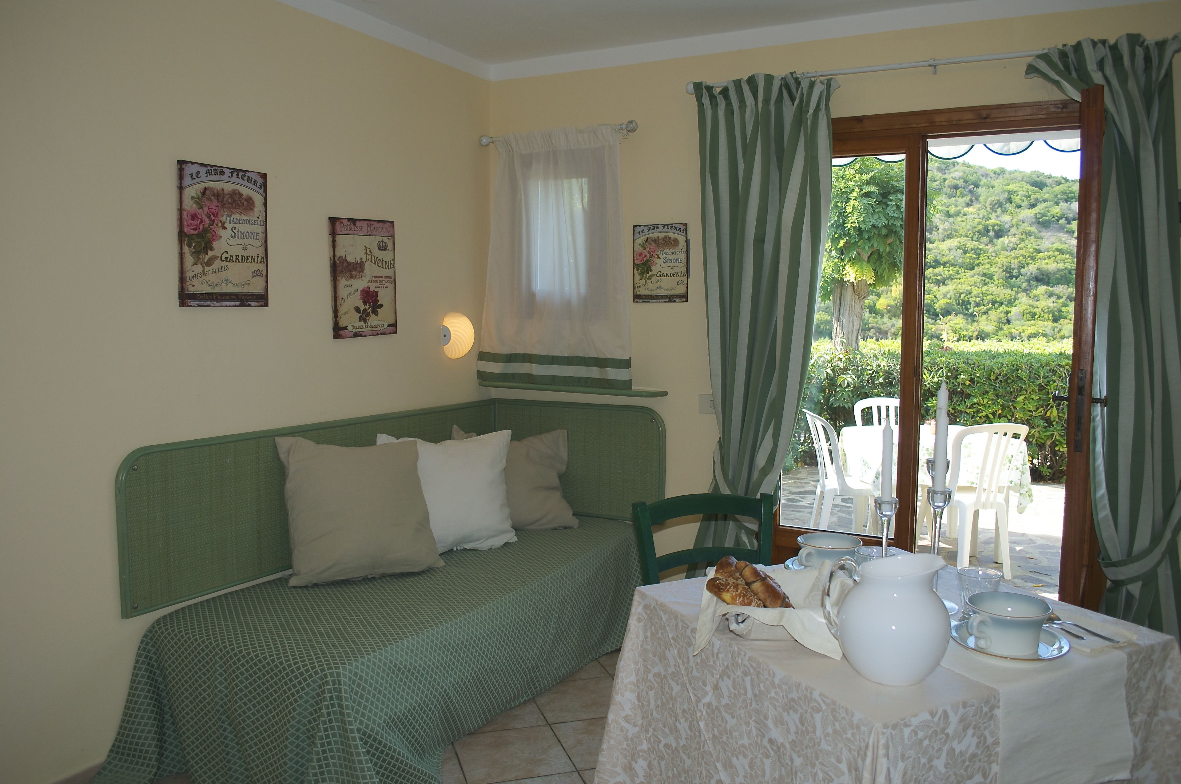 A room with a green sofa, a table with a white tablecloth, and a view of a garden through a window with green curtains.