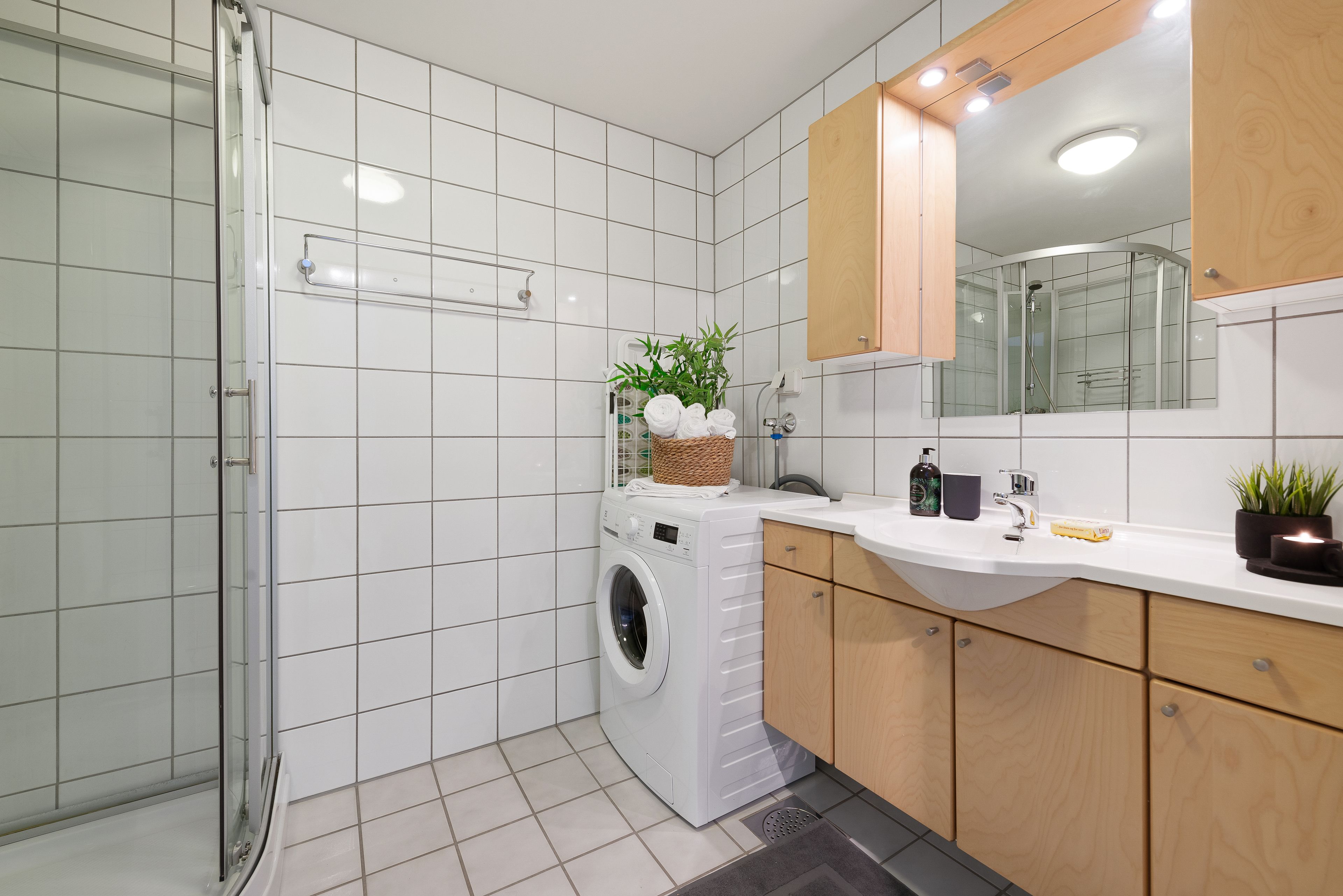 A modern bathroom with a glass shower enclosure, a built-in washing machine, a sink with a mirror, and wooden cabinets.