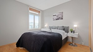 A bedroom with a large bed, a nightstand with a lamp, a small plant, and a city view through the window. - Bjørvika Apartments, Solsiden Area, Trondheim city center (Trondheim)