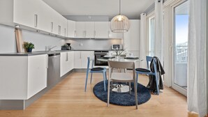 A modern kitchen with white cabinets, a dining area with a round table and chairs, and a view through large windows. - Bjørvika Apartments, Solsiden Area, Trondheim city center (Trondheim)