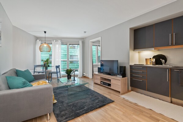Una sala de estar moderna con sofá, mesa de centro y televisor. Hay una zona de cocina con armarios y una mesa de comedor.
