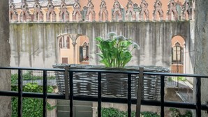 Een balkon met een rieten mand waarin een plant staat, met uitzicht op een historisch gebouw met versierde architectuur en een gietijzeren hekwerk.