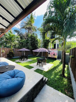 Un patio con mesa y sillas, un sillón de peluche azul y una zona de estar cubierta con sombrillas.