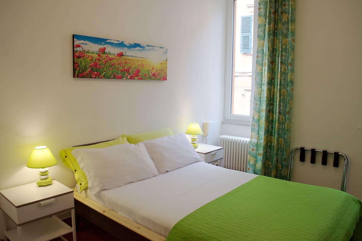 A bedroom with a bed, two bedside tables with lamps, a radiator, and a window with curtains.