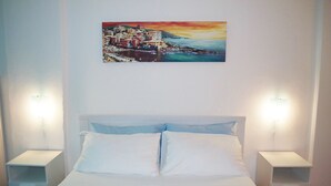 A bedroom with a bed, white nightstands, and a painting of a coastal town on the wall. - Giramondo Room and Breakfast (Rapallo)
