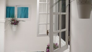 A room with a white wall, a window with blue curtains, a hanging flower pot, and two paintings. - Giramondo Room and Breakfast (Rapallo)