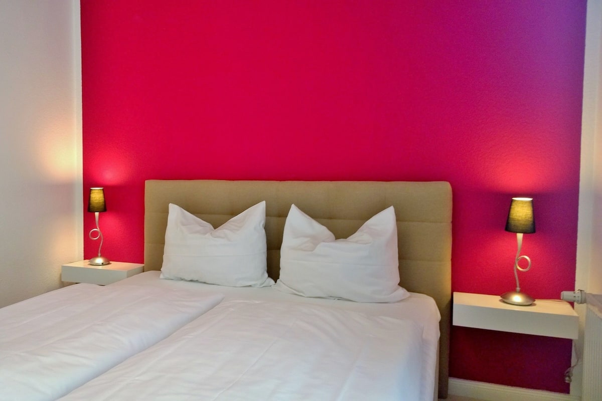 Una cama doble con sábanas blancas, un cabecero color beige y dos lámparas de noche.