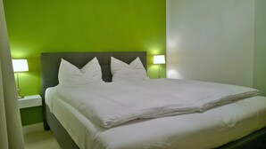 Ein ordentlich bezogenes Bett mit weißen Leinen und Kissen in einem Zimmer mit grünen Wänden und einem grauen Kopfteil. Auf beiden Seiten des Bettes stehen jeweils Nachttischlampen.