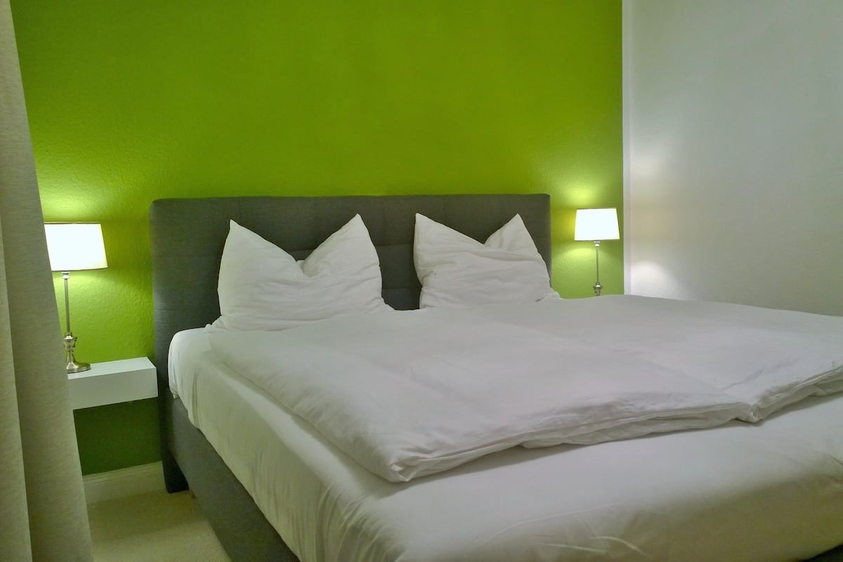 Una cama bien hecha con sábanas y almohadas blancas en una habitación con paredes verdes y un cabecero gris. Hay dos lámparas de noche a cada lado de la cama.