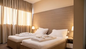 Habitación de hotel con dos camas, cada una con una almohada y una toalla doblada. Hay lámparas de noche y una ventana con cortinas.