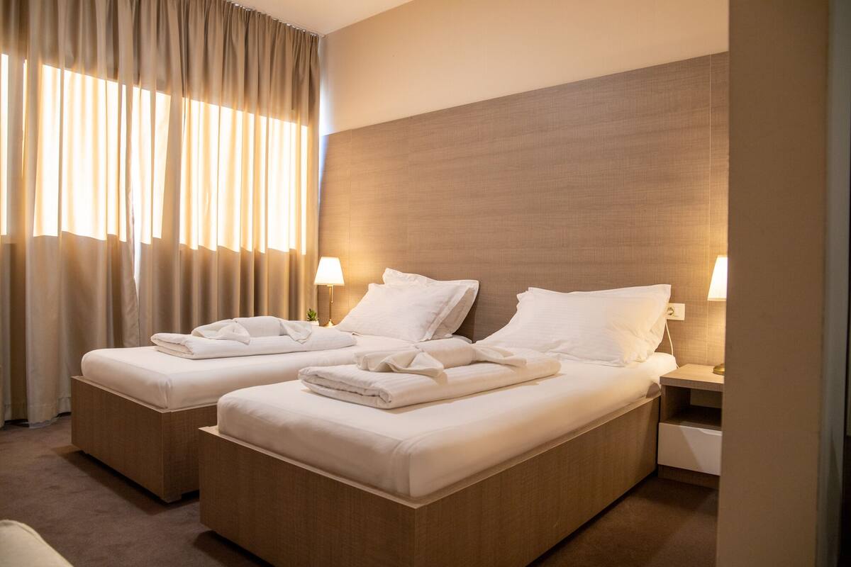 A hotel room with two beds, each with a pillow and a folded towel. There are bedside lamps and a window with curtains.