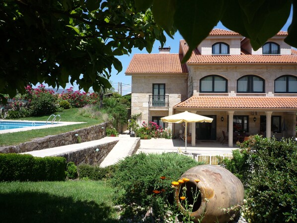A large house with a swimming pool, a patio with an umbrella, and a garden with flowers and a large decorative pot.