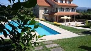 Una casa de dos pisos con piscina, un patio cubierto con mobiliario exterior y fachada de piedra.