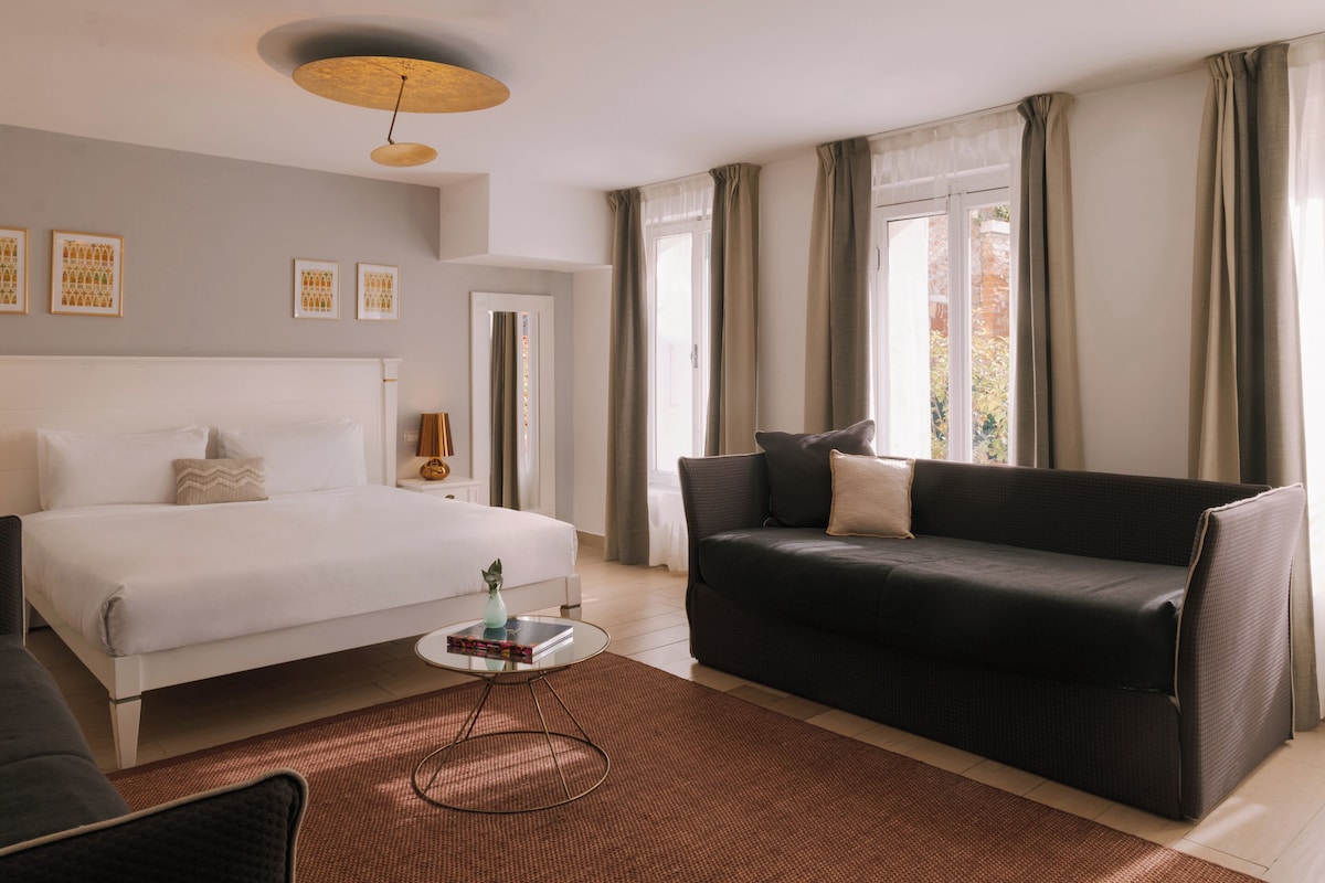 Une chambre d’hôtel avec un lit, un canapé de couleur sombre, une petite table avec un vase et une grande fenêtre avec des rideaux.