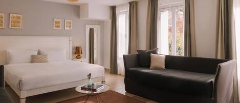 Una habitación de hotel con cama, sofá oscuro, una mesita con un florero y un ventanal con cortinas.