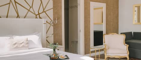 Une chambre d’hôtel avec un grand lit, une petite table sur laquelle se trouvent un vase et des pâtisseries, un fauteuil à la structure dorée et un canapé vert.