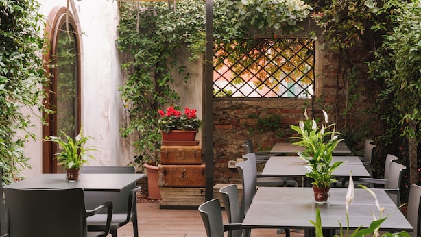 Un patio al aire libre con mesas y sillas, rodeado de follaje y un muro de ladrillo.