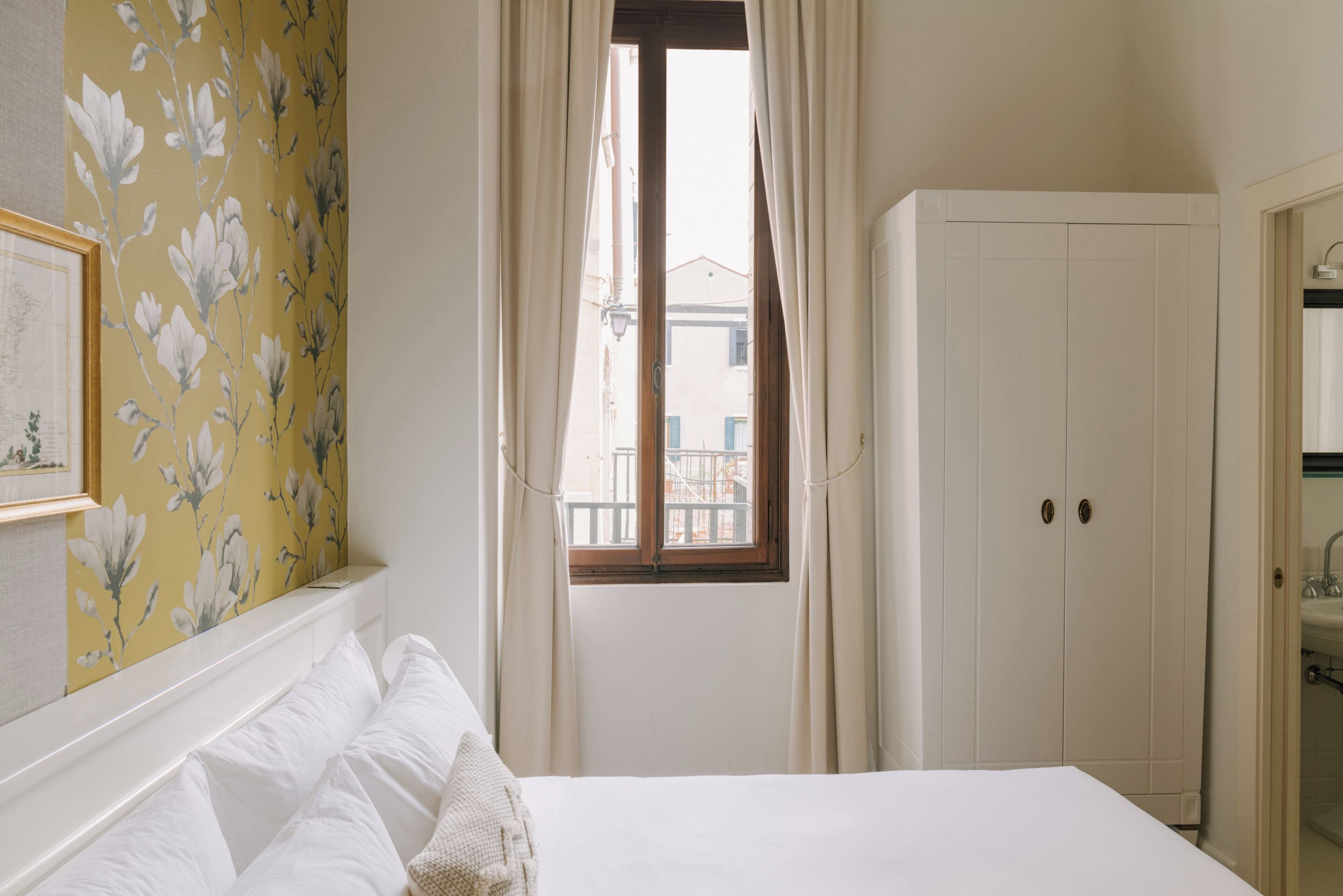 Un dormitorio con cama, ventana con cortinas, armario y pared con papel tapiz floral.