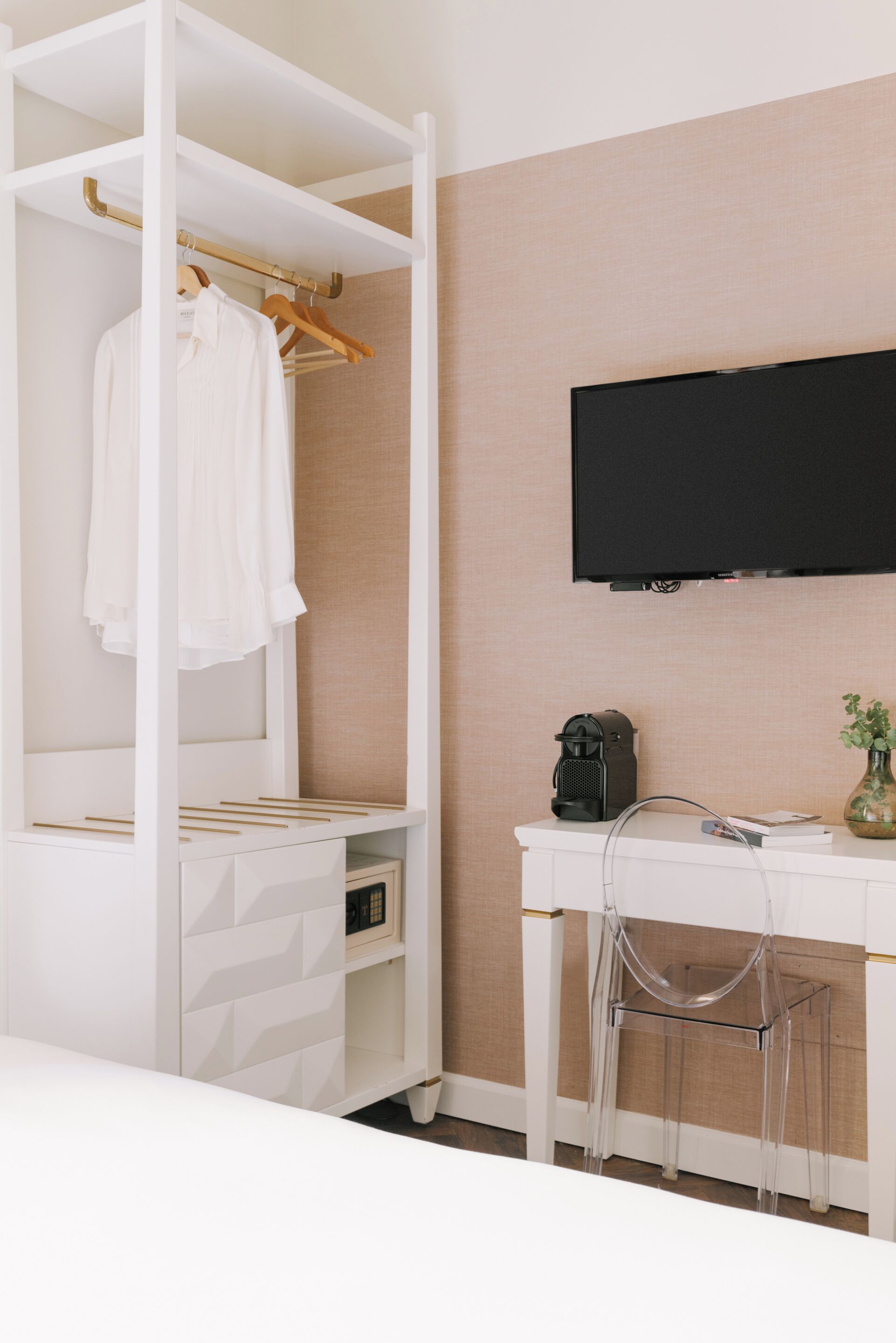 A bedroom with a white wardrobe, a desk with a chair, a coffee maker, and a TV on the wall.