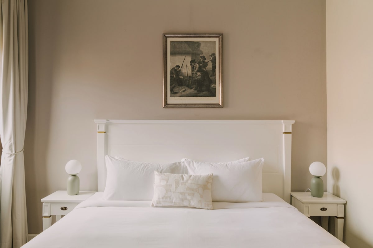 Un lit bien fait, avec du linge de lit blanc et deux oreillers, une œuvre d’art encadrée au-dessus du lit, et deux tables de chevet avec des lampes.