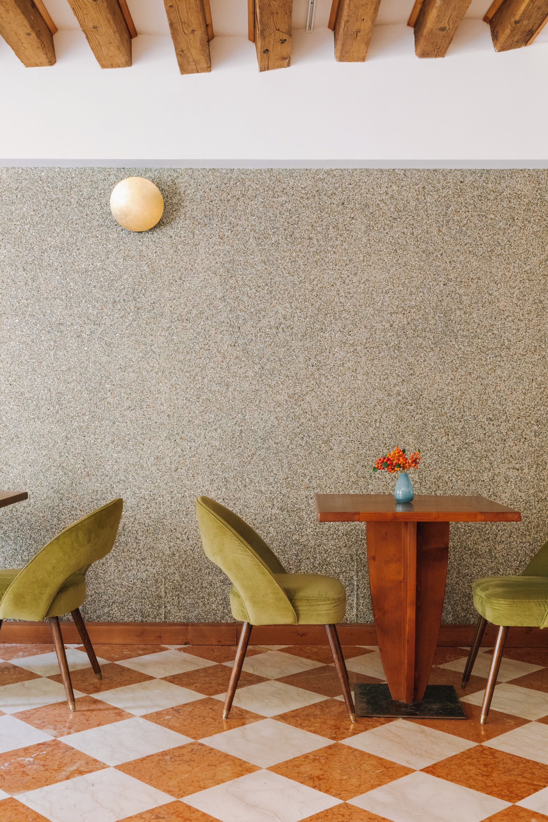 A dining area with a checkered floor, two green chairs, a wooden table with a vase of flowers, and a textured wall.
