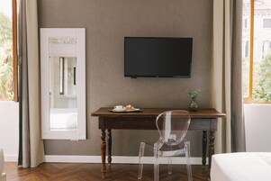 Una stanza con una scrivania in legno, una sedia trasparente, una televisione a schermo piatto fissata al muro, un piccolo vaso con una pianta e una finestra con tende.