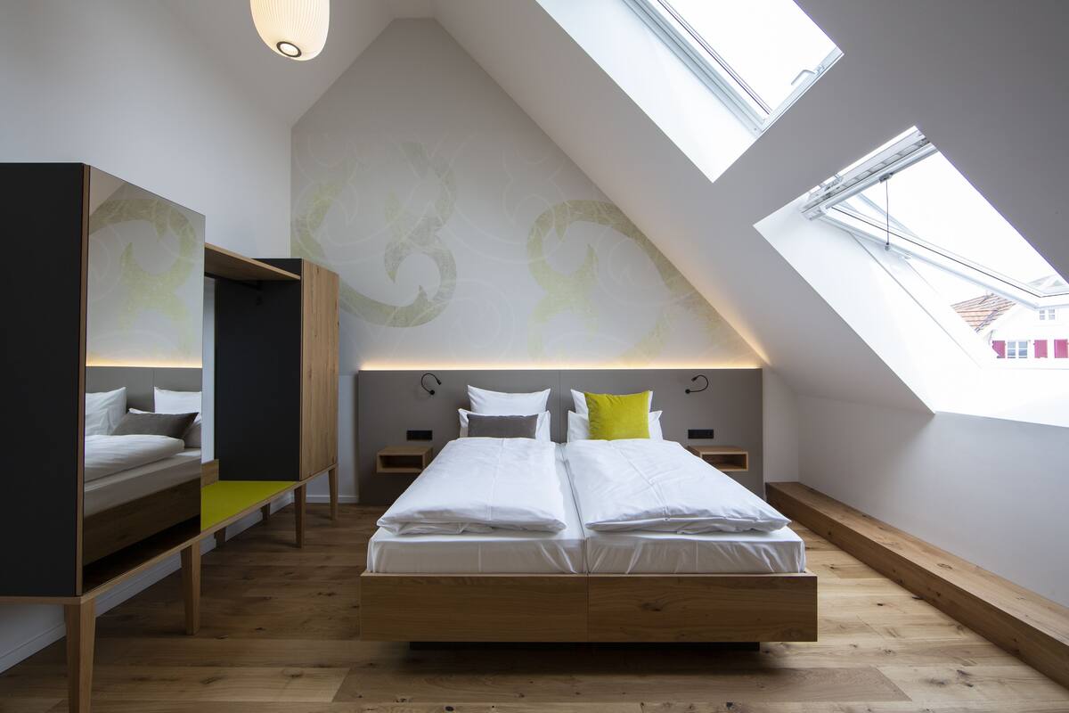 Ein modernes Schlafzimmer mit schrägen Wänden, einem Dachfenster, einem Bett mit weißer Bettwäsche und einer Bank mit Kissen.
