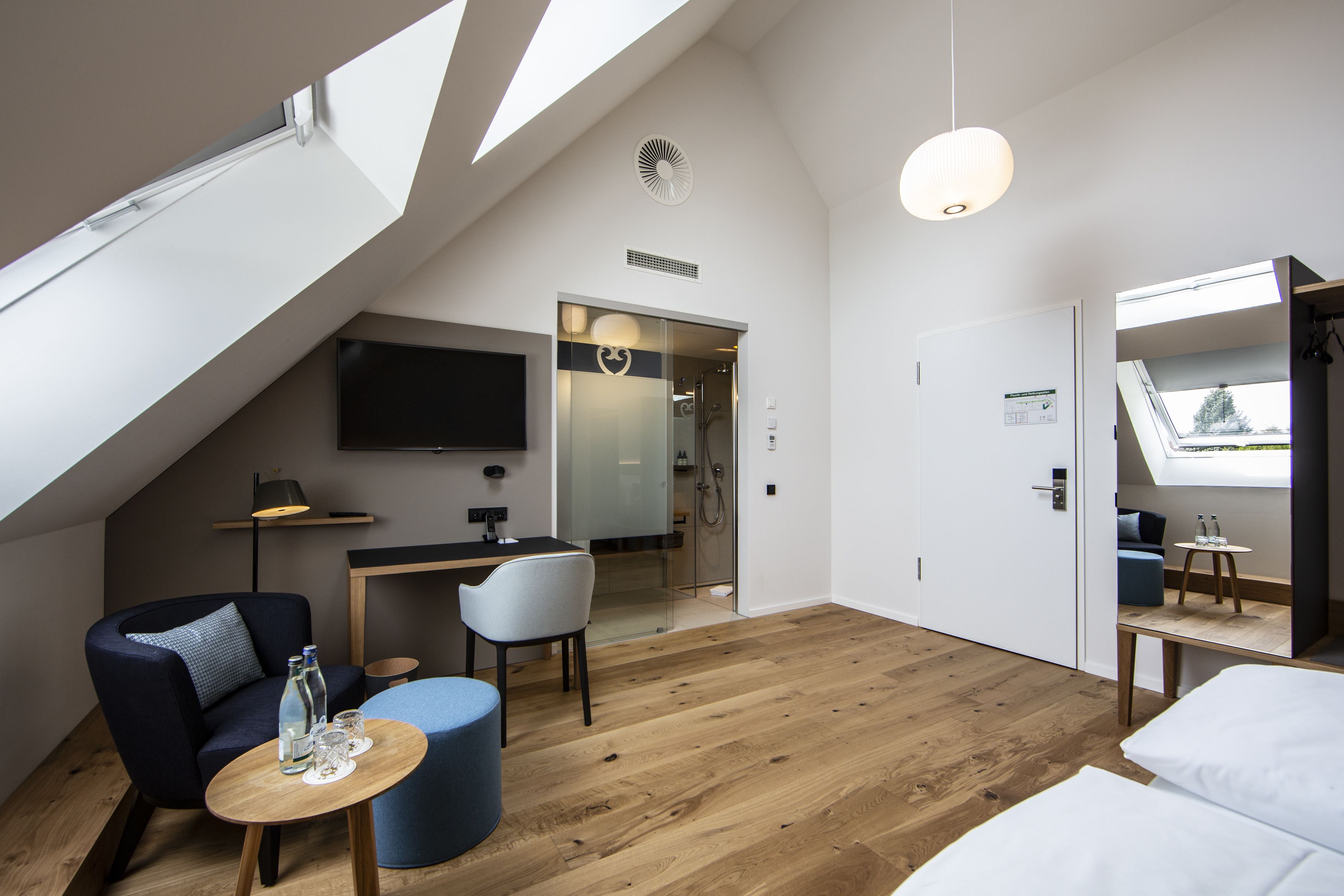 Una habitación de hotel moderna con techo inclinado, un televisor de pantalla plana montado en la pared, un escritorio con una silla, una pequeña mesa redonda con una botella y vasos, y una cama con ropa blanca.