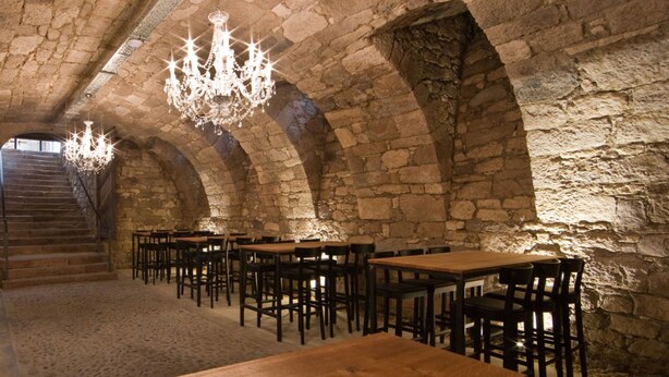 Une pièce aux murs de pierre, avec des tables et des chaises en bois, un escalier et des lustres.
