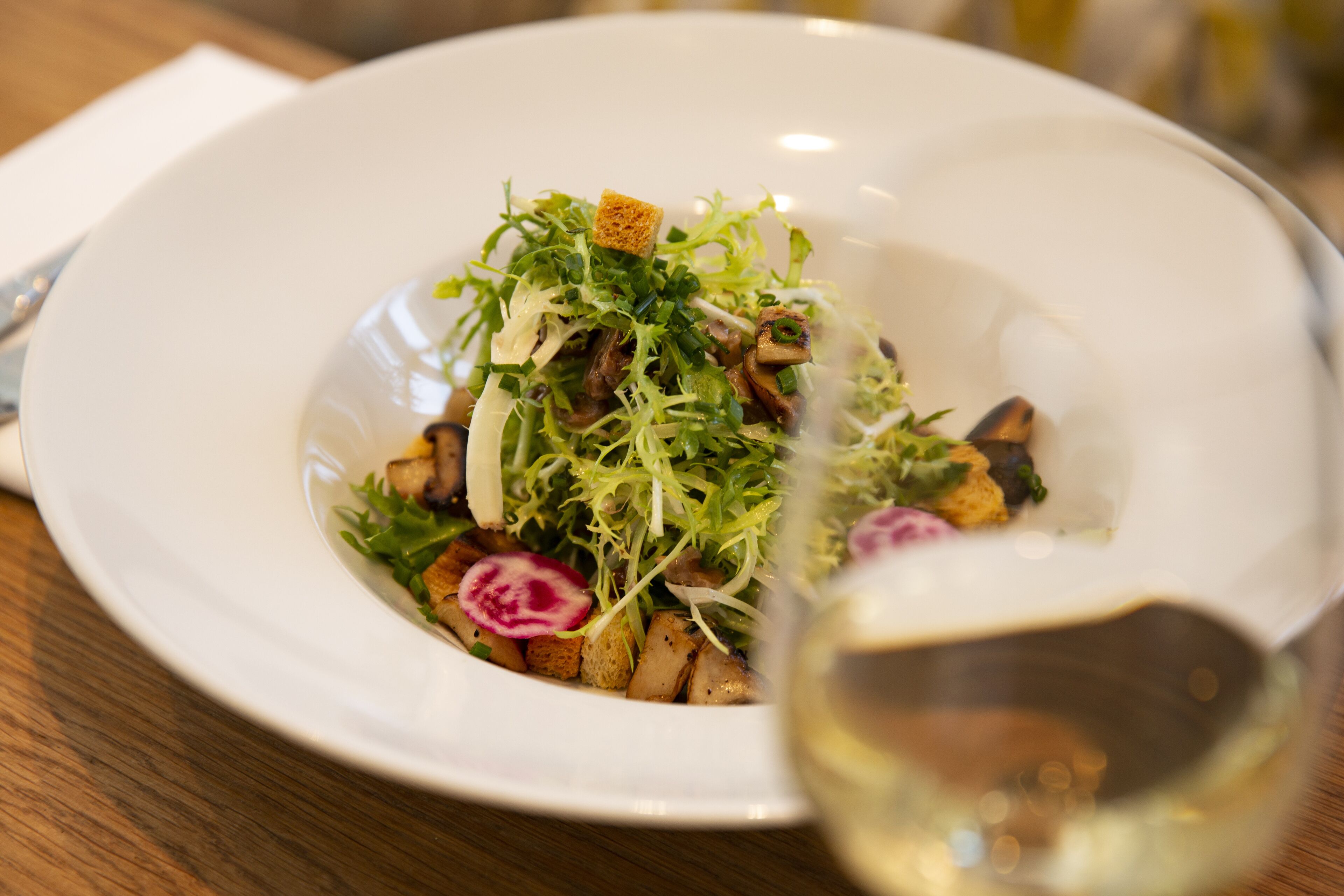 A white plate with a salad, croutons, and radish slices on a wooden table.