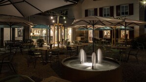 Un espace extérieur aménagé en terrasse, avec des tables et des chaises, des parasols et une fontaine centrale.