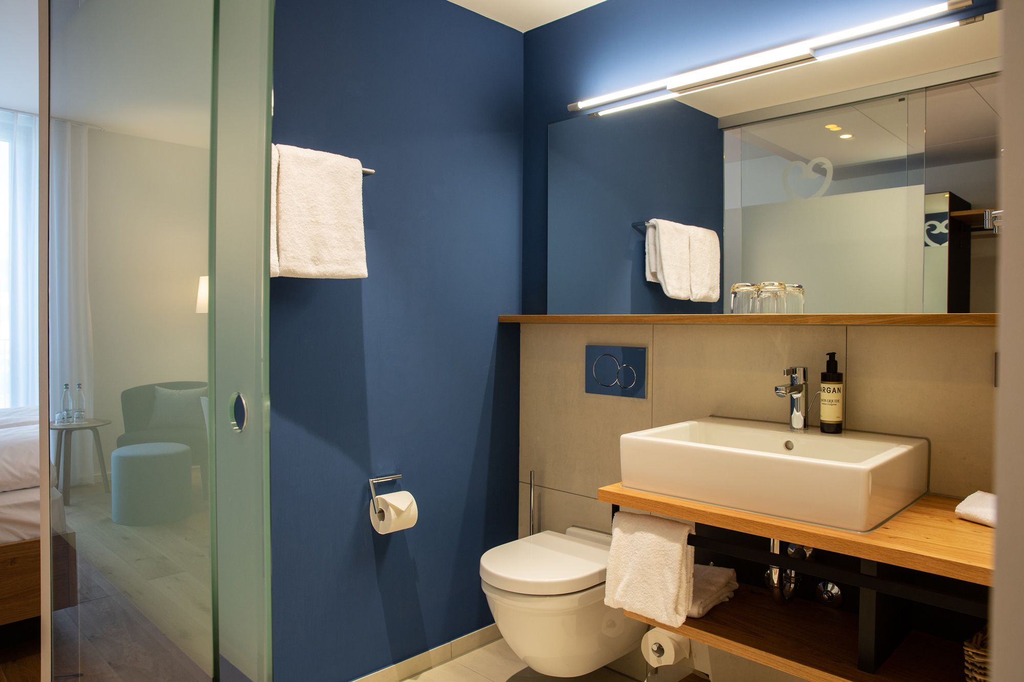 A modern bathroom with a blue wall, a glass shower enclosure, a white toilet, and a wooden vanity with a mirror.