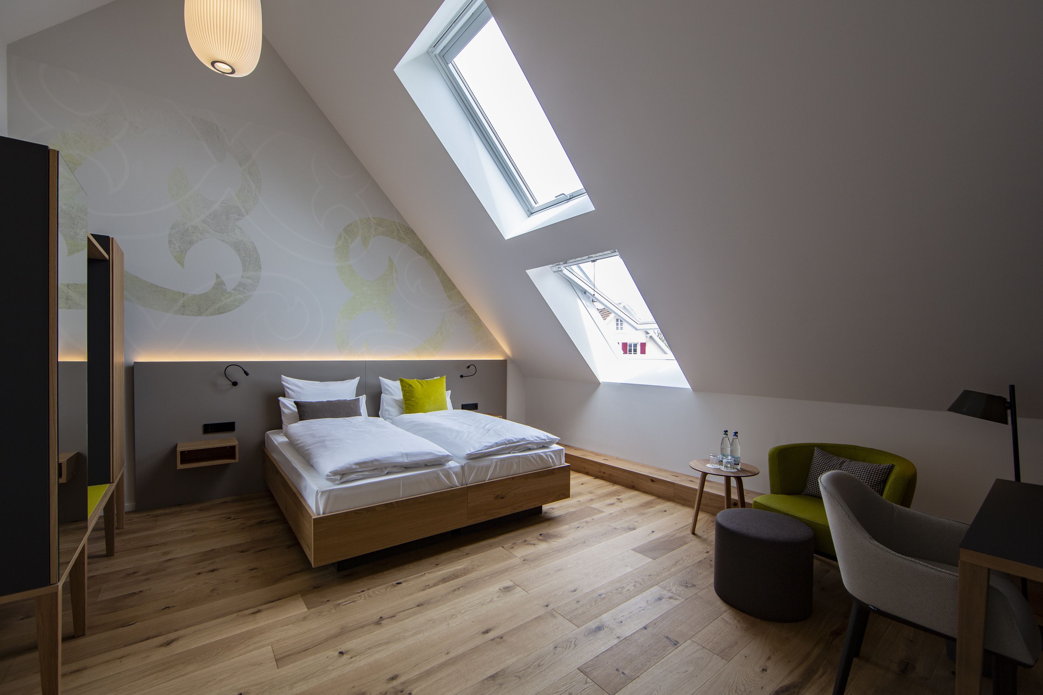 Ett modernt sovrum med ett tak i loftstil, en säng med vita sängkläder, en grön fåtölj och ett litet bord med två flaskor.