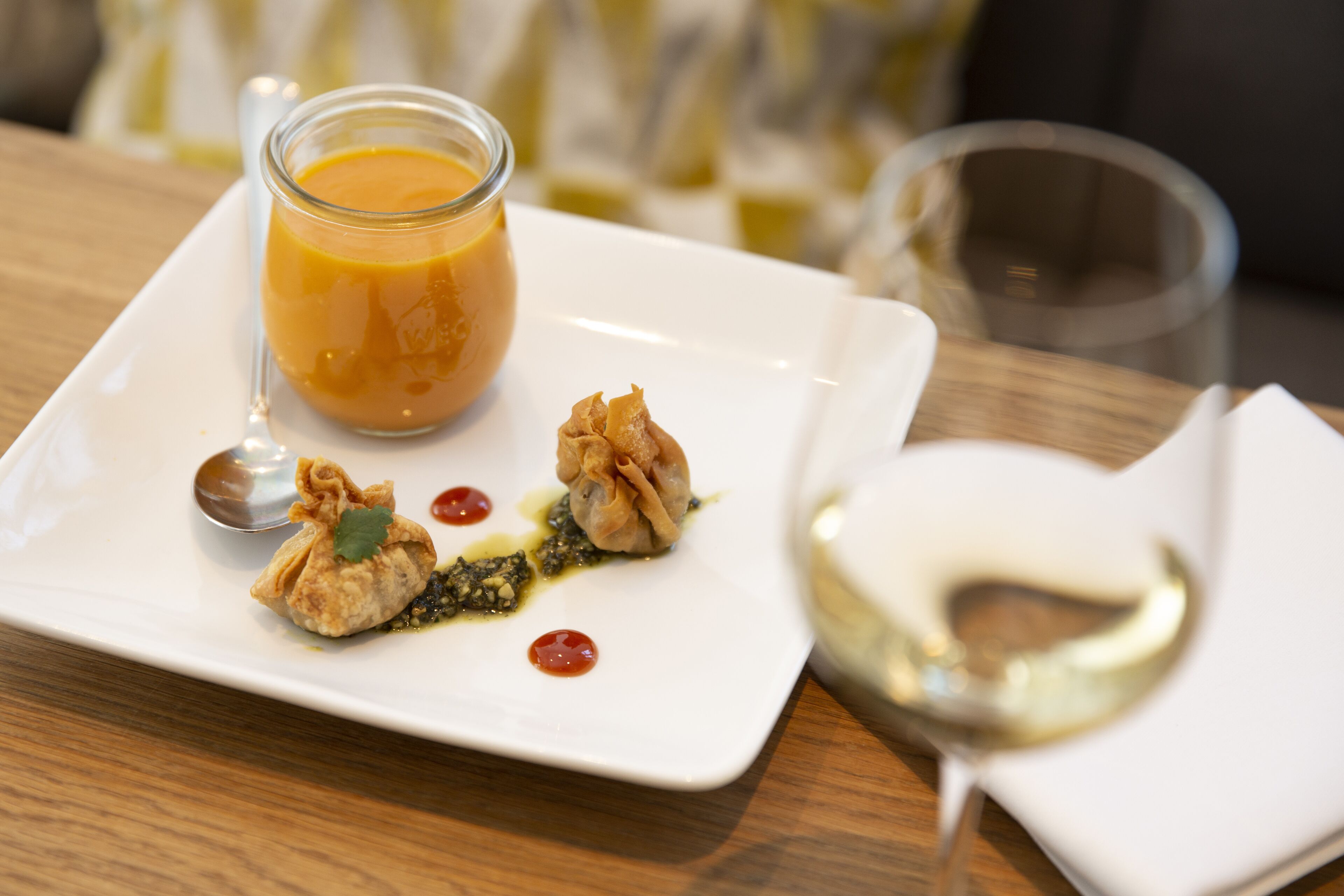 A white square plate with a glass of orange juice, two dumplings, a spoon, and a small dish of sauce.