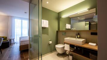 A modern bathroom with a glass shower enclosure, a toilet, a sink with a mirror, and a bedside table with a chair.