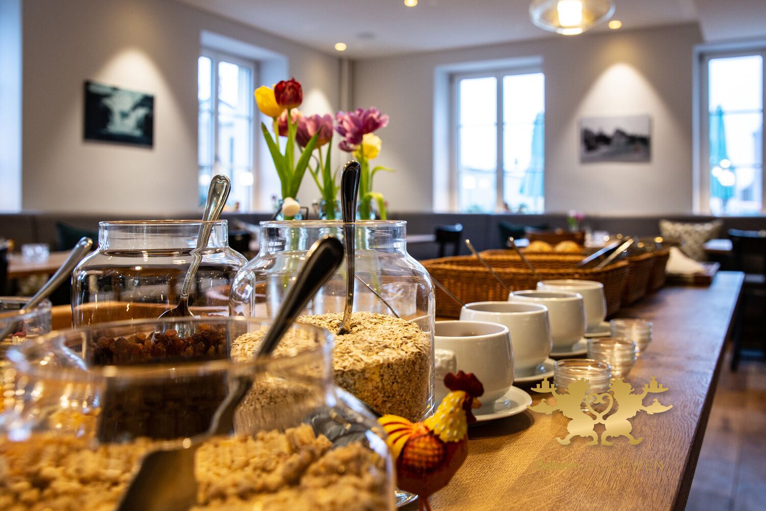 A buffet setup with various food items and utensils, a vase with flowers, and a view of the room with windows and artwork.