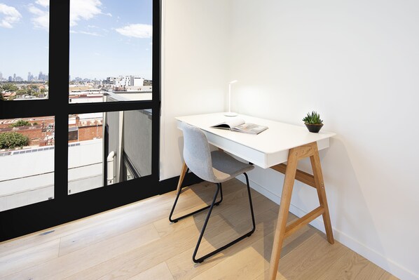 A modern office space with a white desk, a grey chair, a small potted plant, and a city view through large windows. - Artel Apartment Hotel Melbourne (Brunswick)