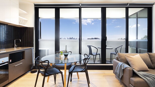 A modern living room with a glass table and chairs, a sofa, and a city view through large windows. - Artel Apartment Hotel Melbourne (Brunswick)