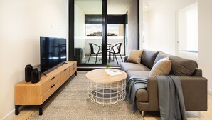 A modern living room with a sofa, a TV, a wooden cabinet, a round coffee table, and a view through a glass door to an outdoor area with chairs. - Artel Apartment Hotel Melbourne (Brunswick)