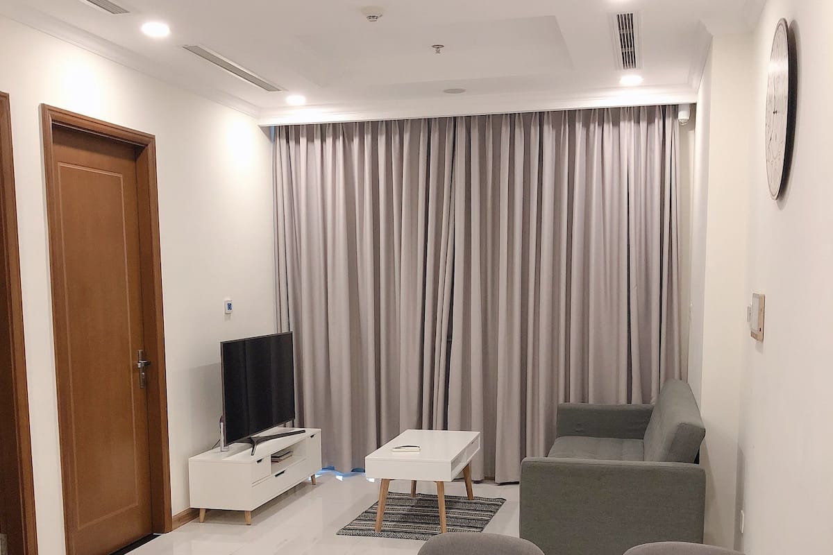 Ein modernes Wohnzimmer mit einem weißen Sofa, einem Esstisch mit Stühlen, einem Fernseher auf einem Schrank und einem großen Fenster mit Vorhängen.