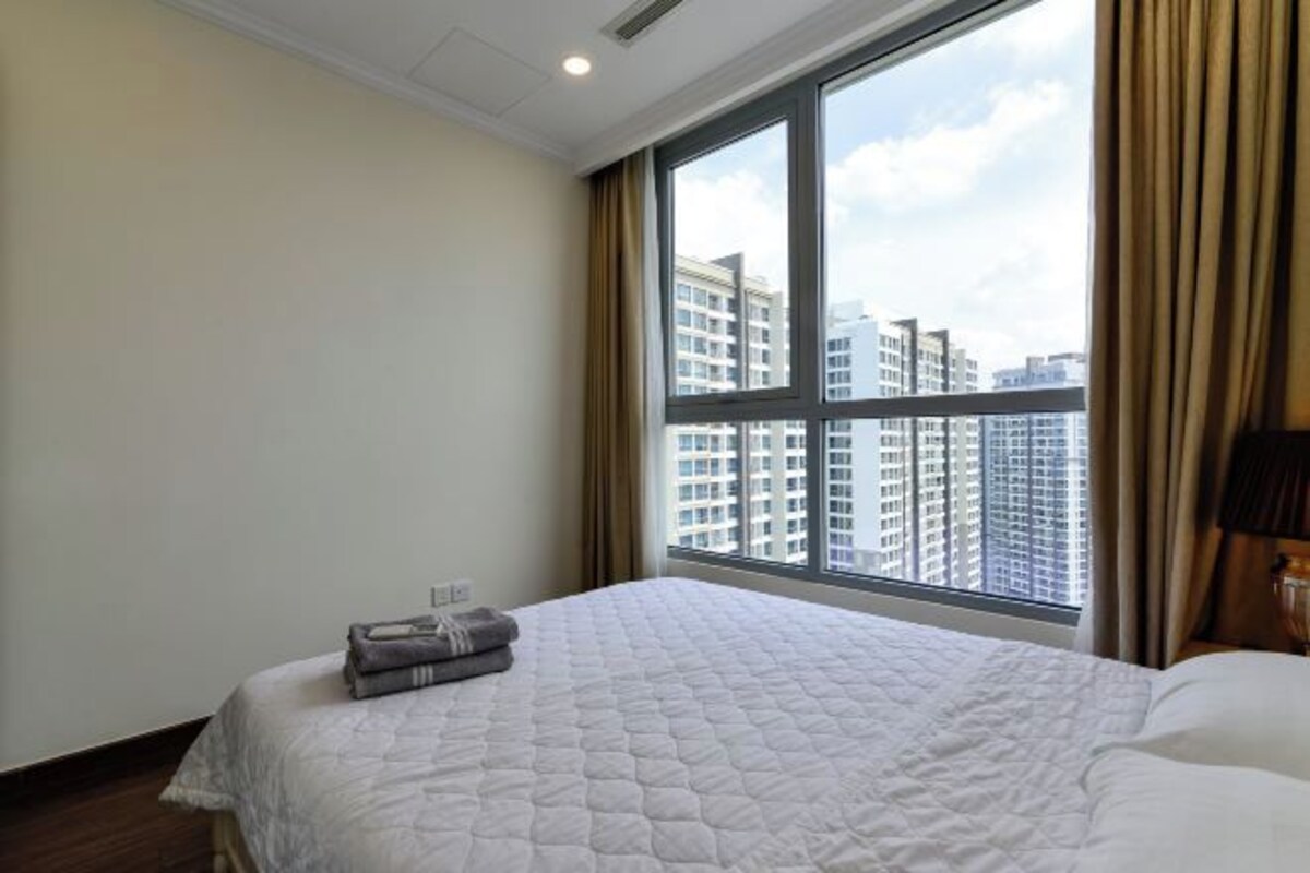 A hotel room with a large window, a bed with a white quilt, and a view of high-rise buildings.