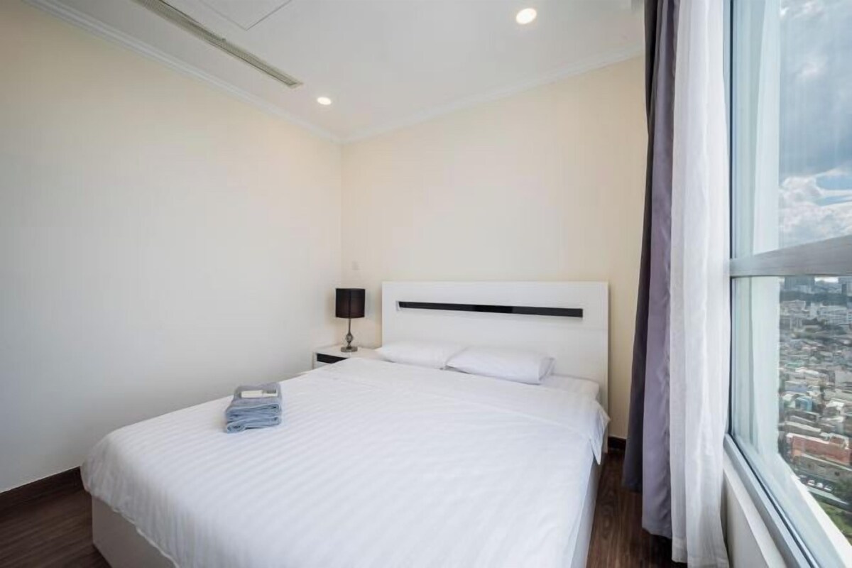 Habitación de hotel con una cama grande, una mesita de noche, una lámpara y una ventana con vistas a la ciudad.