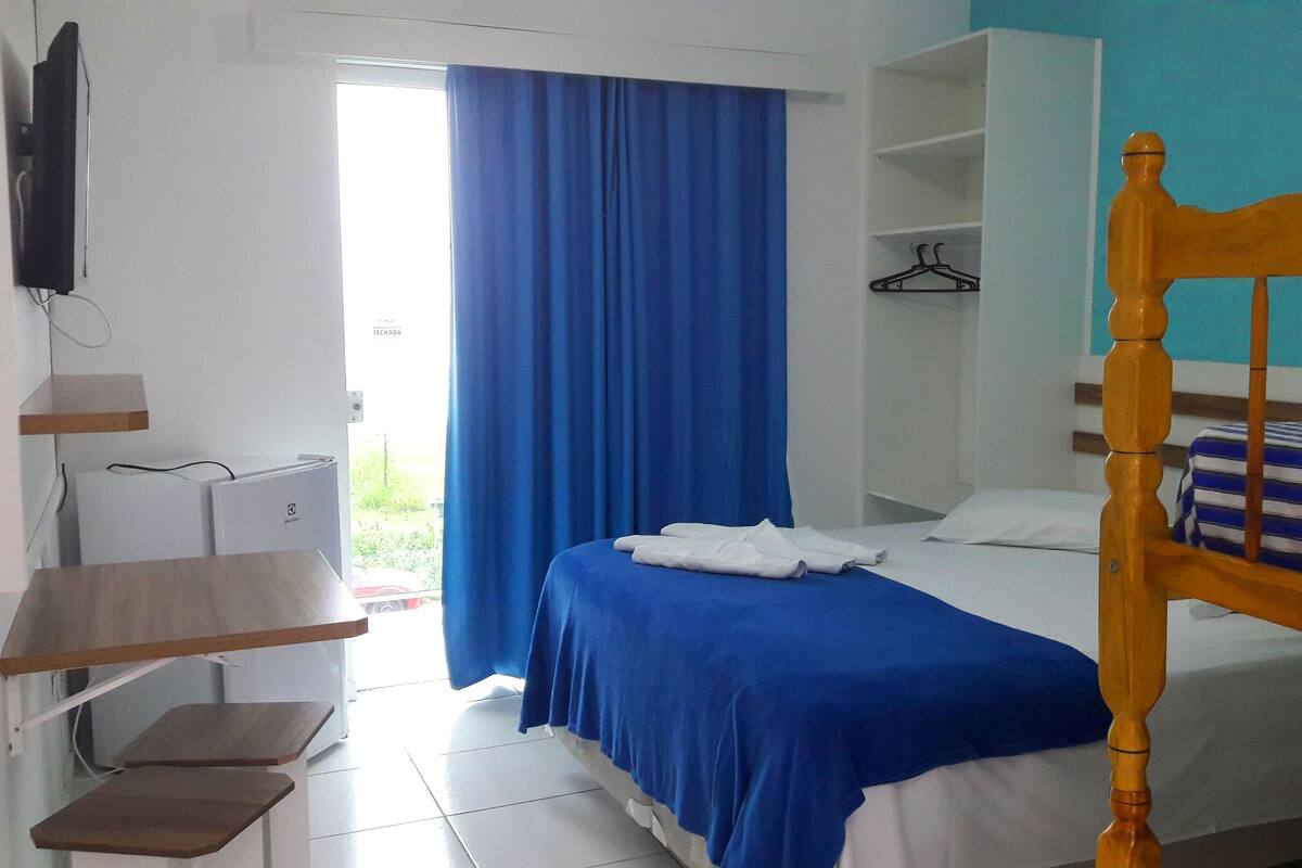 A bedroom with a single bed, a wooden headboard, a blue curtain, a TV mounted on the wall, and a small desk with a chair.